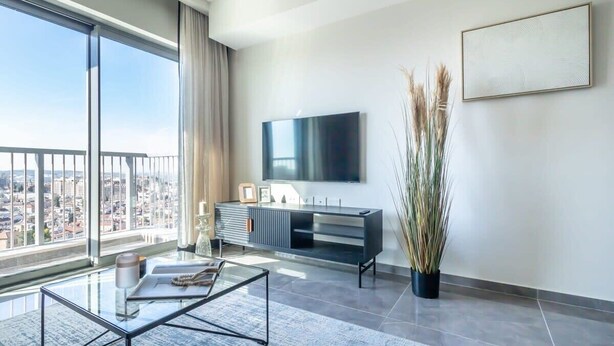 A modern living room with a large window, a flat-screen TV, a minimalist console table, and a tall plant. - Fantastic 1 Bedroom In City Center - isrentals (Jerusalem)