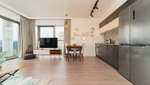 A modern living room with a dining area, a large flat-screen TV, and a kitchen with a stainless steel refrigerator. - 1 Bedroom Savyon View - Isrentals (Jerusalem)