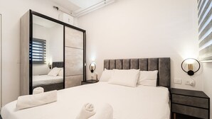 A modern bedroom with a large bed, a wardrobe with a mirror, bedside lamps, and a nightstand. - 1 Bedroom Savyon View - Isrentals (Jerusalem)