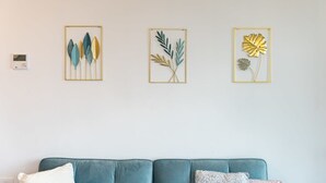A room with a teal sofa, three framed botanical artworks on the wall, and a thermostat on the left. - 1 Bedroom Savyon View - Isrentals (Jerusalem)