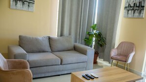 A living room with a grey sofa, two pink chairs, a wooden coffee table with remote controls, a potted plant, and artwork on the walls. - The Backyard Lodge (Mpingu)