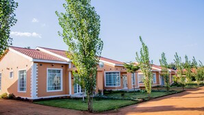 A row of modern, single-story buildings with large windows and a red-tiled roof, surrounded by well-maintained lawns and trees. - The Backyard Lodge (Mpingu)
