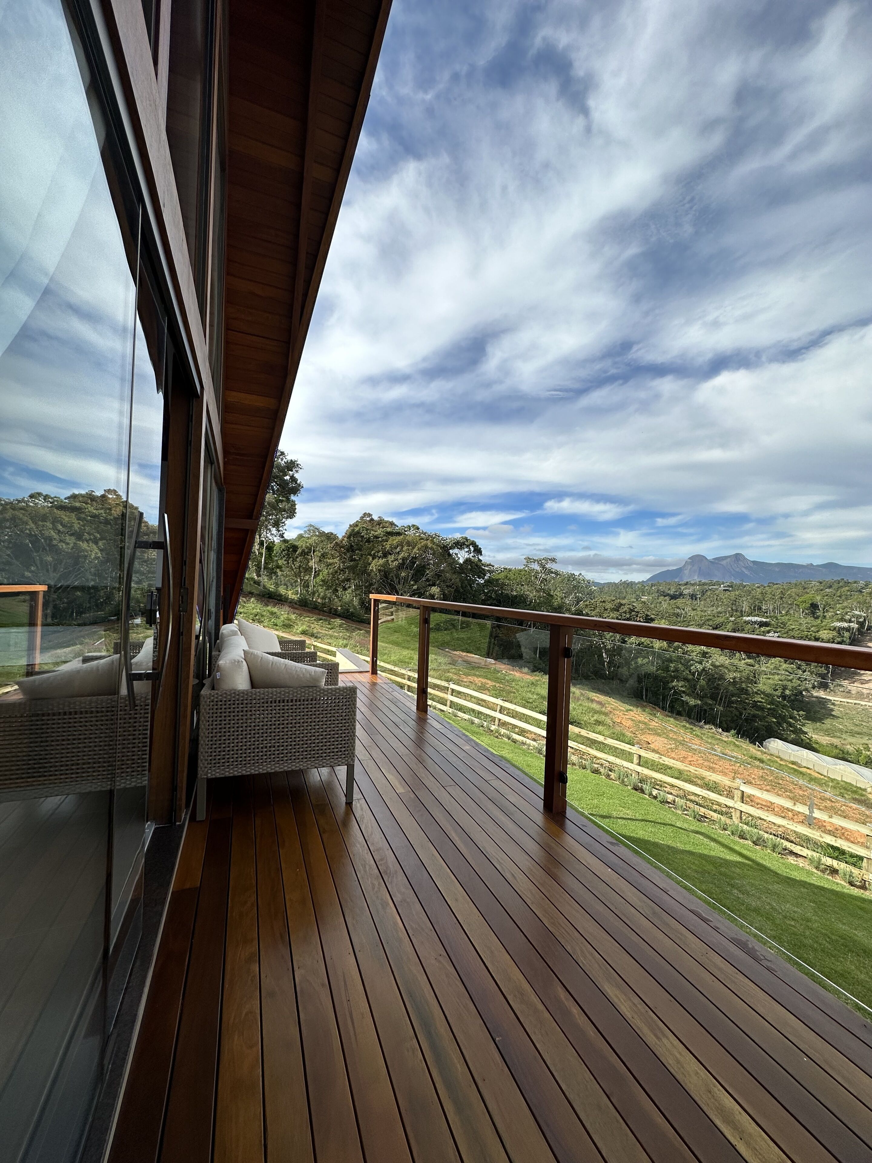 A wooden deck with a glass wall, a wicker sofa, and a view of a green landscape with trees and mountains.