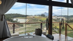 A large, modern bathroom with a bathtub, a view of a scenic landscape through large windows, and a wooden deck outside. - Chalés Laguna Alto Viçosa (Venda Nova Do Imigrante)