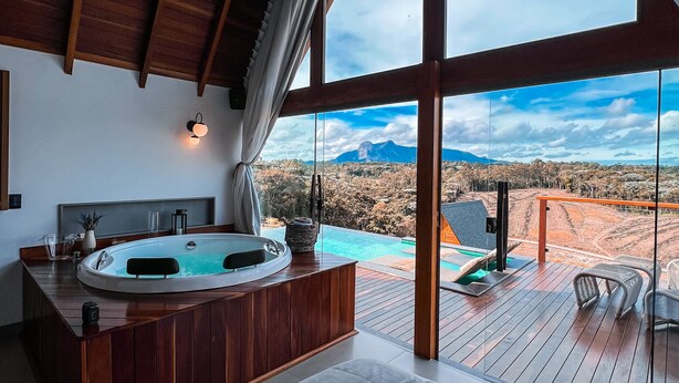 A modern bathroom with a wooden vanity, a large bathtub, and a view of a mountainous landscape through large windows. - Chalés Laguna Alto Viçosa (Venda Nova Do Imigrante)