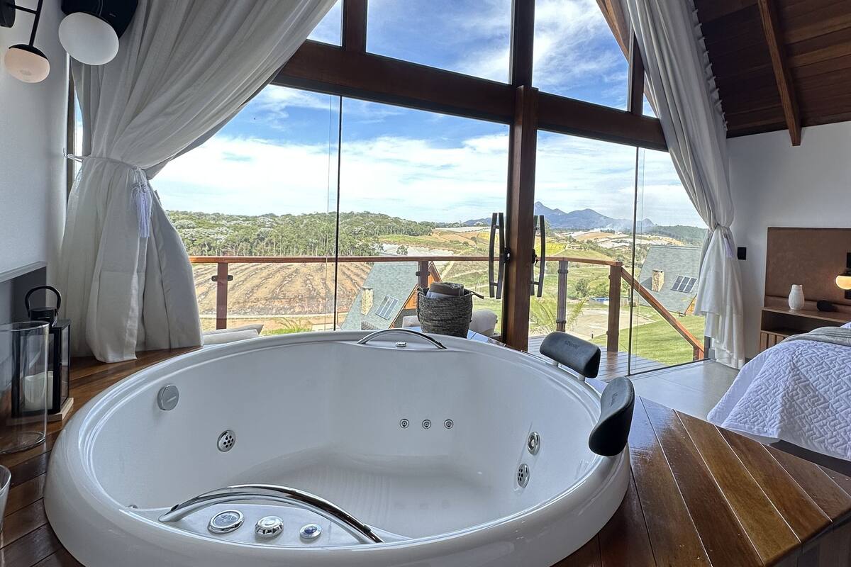 A modern room with a large window, a hot tub, wooden flooring, and a bed.