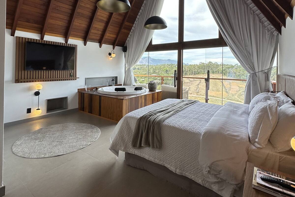 A bedroom with a large bed, a wooden ceiling, a TV mounted on the wall, and a view of the landscape through a window.
