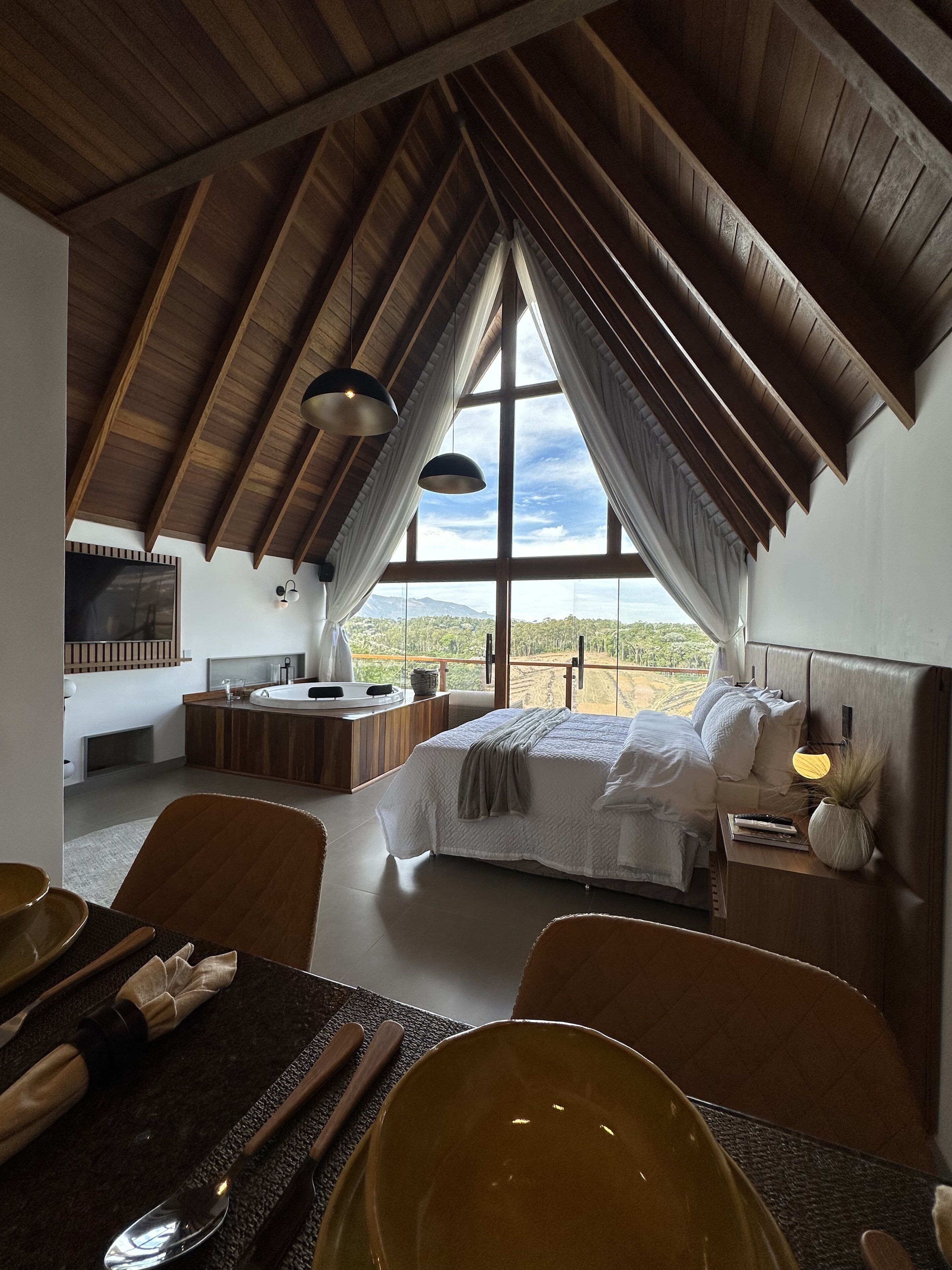 Ein Schlafzimmer mit großem Fenster, Holzbalkendecke, einem Bett, Nachttischen und Blick auf die Landschaft.