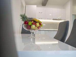A fruit bowl on a table in a modern kitchen with white cabinets and a gray chair.
