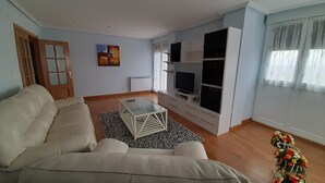 Ein Wohnzimmer mit einem beigen Sofa, einem Glastisch, einem Fernseher auf einer weißen Unterhaltungseinschub und einem Heizkörper.