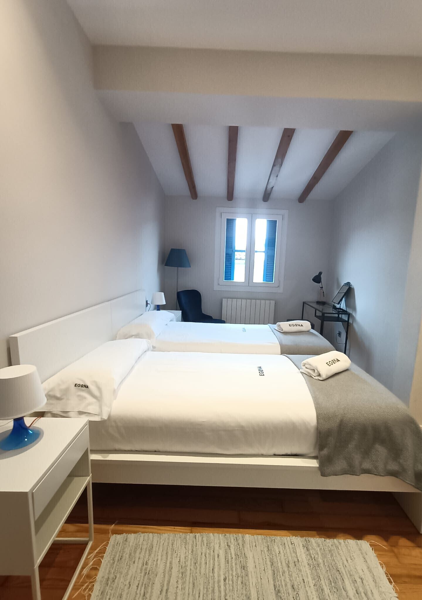 A hotel room with two beds, a desk, a chair, a lamp, a window, and a radiator.