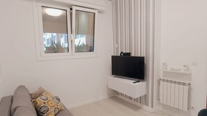 A modern living room with a sofa, a flat-screen TV mounted on a white console, a window with curtains, and a radiator. - Apartment Frida - Nice, 150m from the beach by Egona (Zarautz)