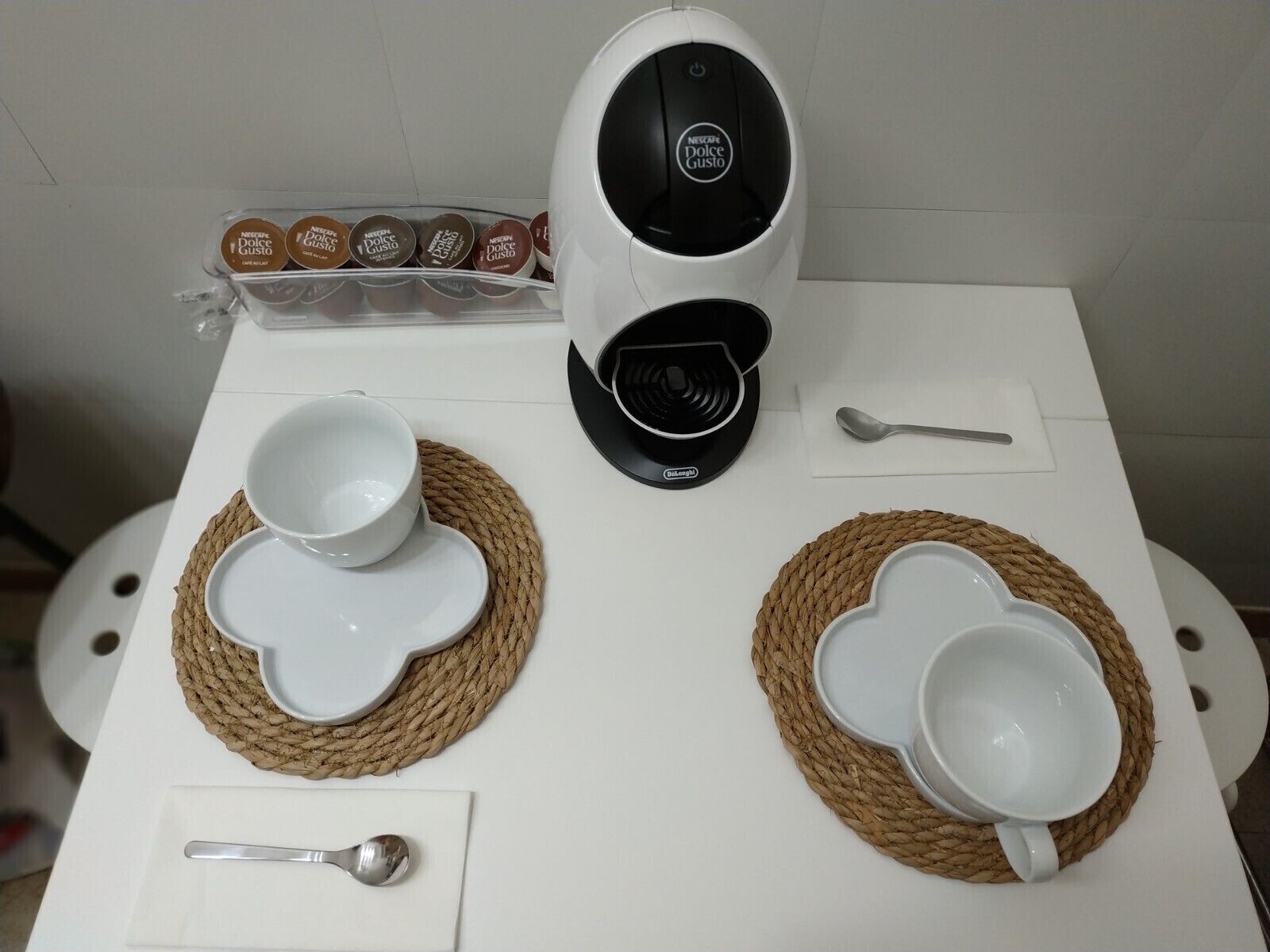 A coffee maker, two cups, a spoon, and a container of Dolce Gusto coffee pods on a table.