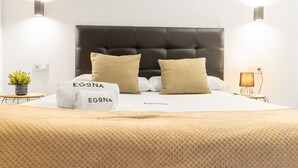 Een net opgemaakt bed met twee kussens, een nachtkastje met een lamp, en een wandlamp.