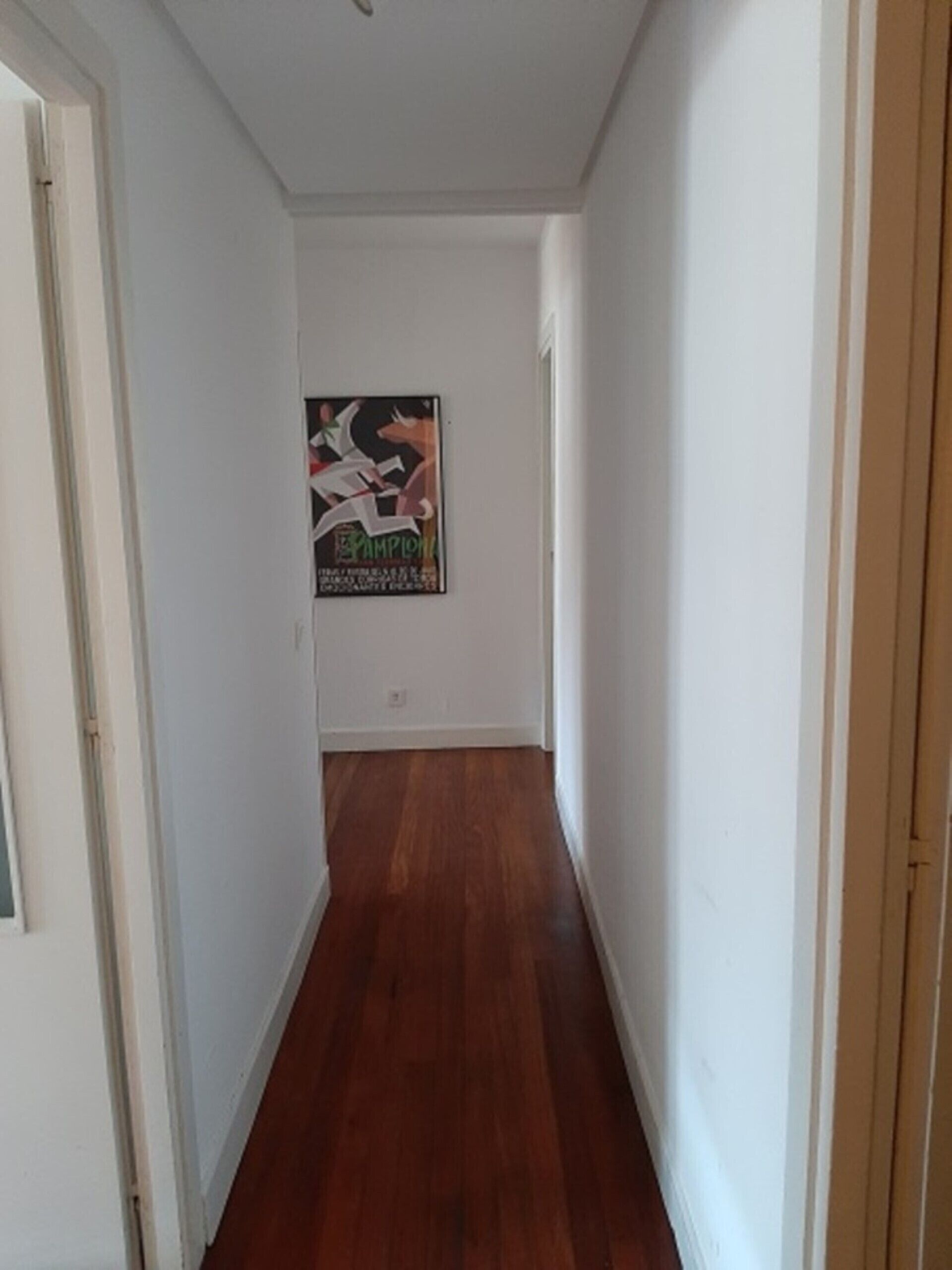Een smalle gang met houten vloer en witte wanden. Er hangt een poster aan de muur en links bevindt zich een deur.