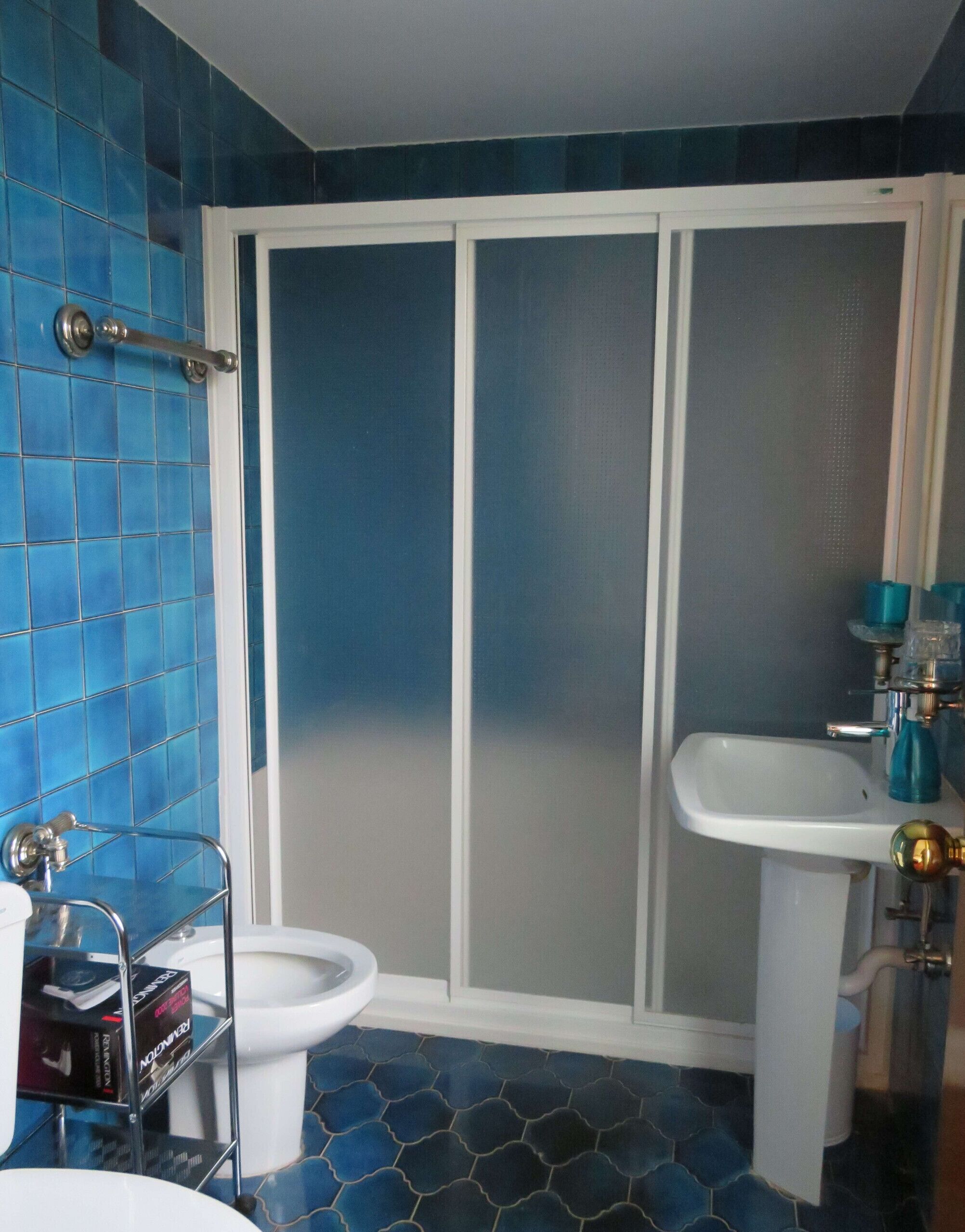 Een badkamer met blauwe betegelde wanden, een wit toilet, een douchecabine en een wastafel met een blauwe kraan.