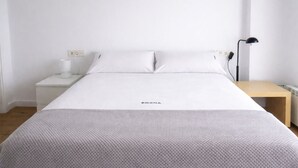 Een netjes opgemaakt bed met een witte dekbedovertrek en kussens, een nachtkastje met een lamp, en een houten nachtkastje.