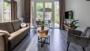 A modern living room with a sofa, armchair, and coffee table. There is a TV on the wall and a view of the outdoors through large glass doors. - Chic Chalet in Hoenderloo (Hoenderloo)