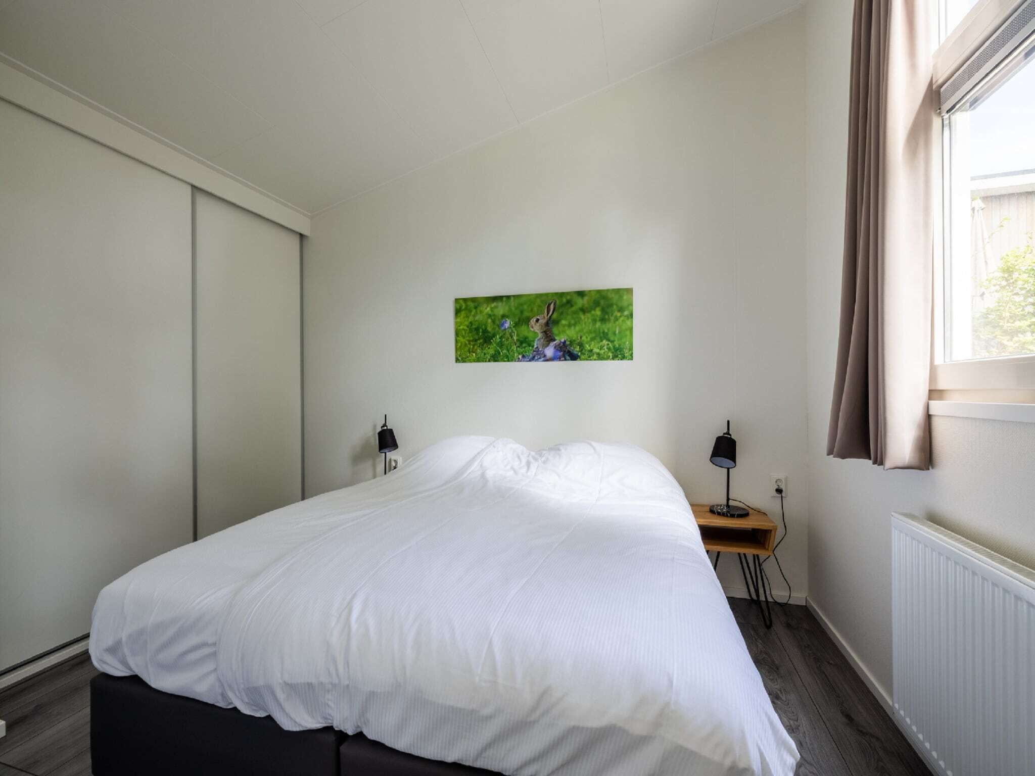 A bedroom with a bed, a nightstand, a lamp, a picture on the wall, and a window with curtains.