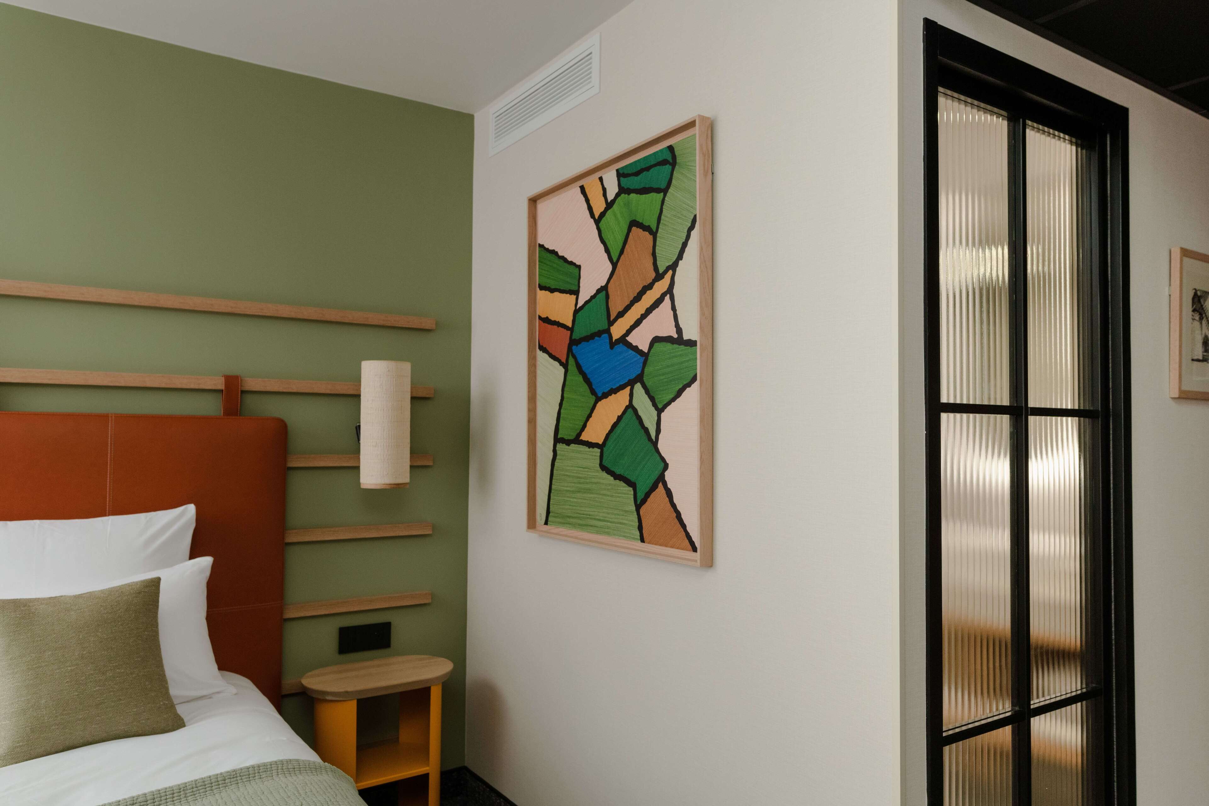 A hotel room with a bed, a wooden headboard, a small wooden nightstand, a wall-mounted artwork, and a window with blinds.