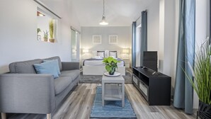A modern living room with a grey sofa, a coffee table with a plant, a TV stand, and a bed in the background. - Basalt House B by Homie (São Vicente)