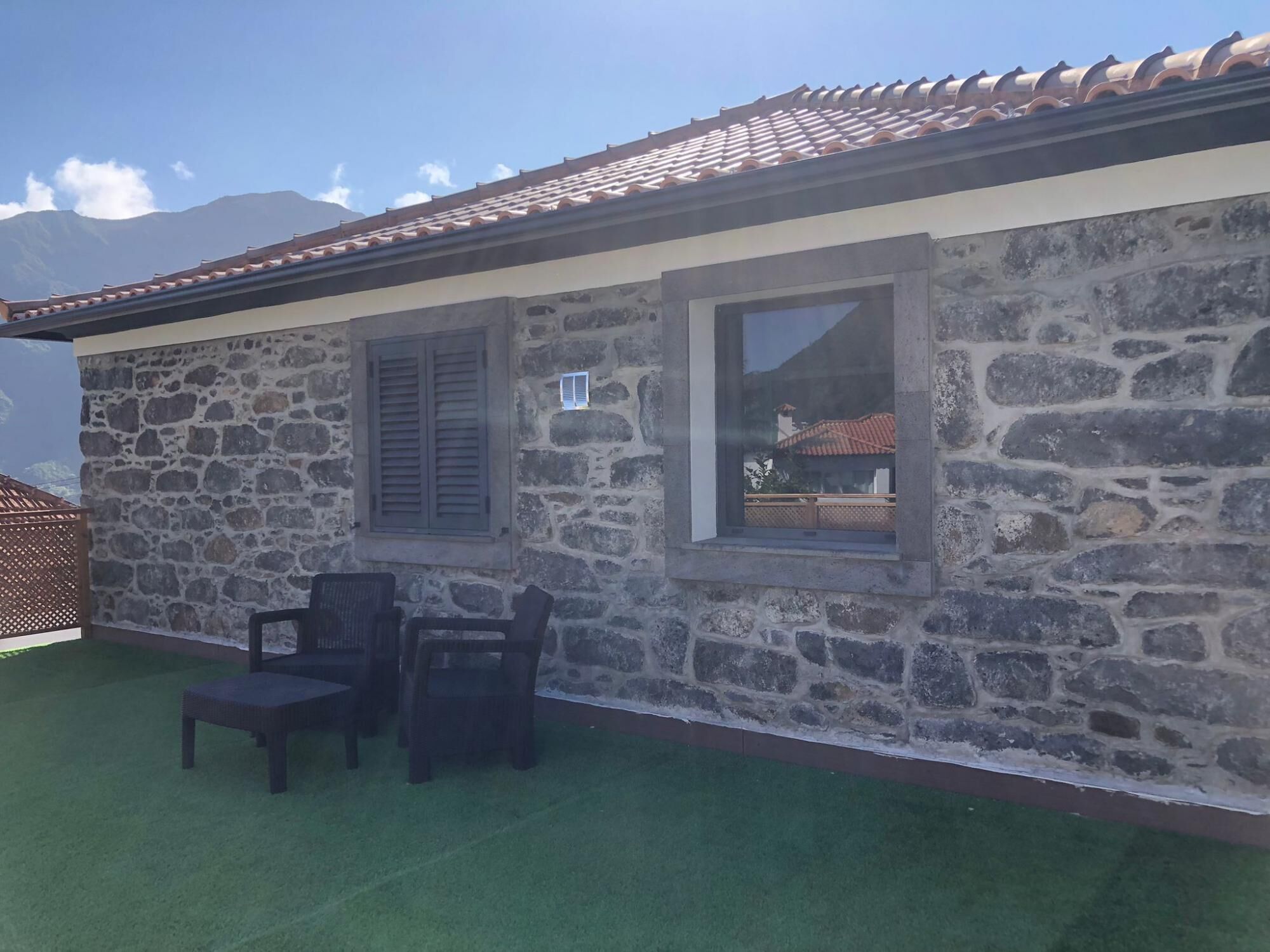 A stone building with a tiled roof, two windows, and a small outdoor seating area with chairs.