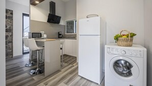 A compact kitchen with a microwave, coffee maker, and a refrigerator. There's a washing machine and a wicker basket with fruits on top. - Basalt House B by Homie (São Vicente)
