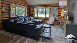 En stuga med träpanel på insidan, med en svart lädersoffa, ett litet bord, en vit fåtölj, en takfläkt och en tv som står på ett skåp.