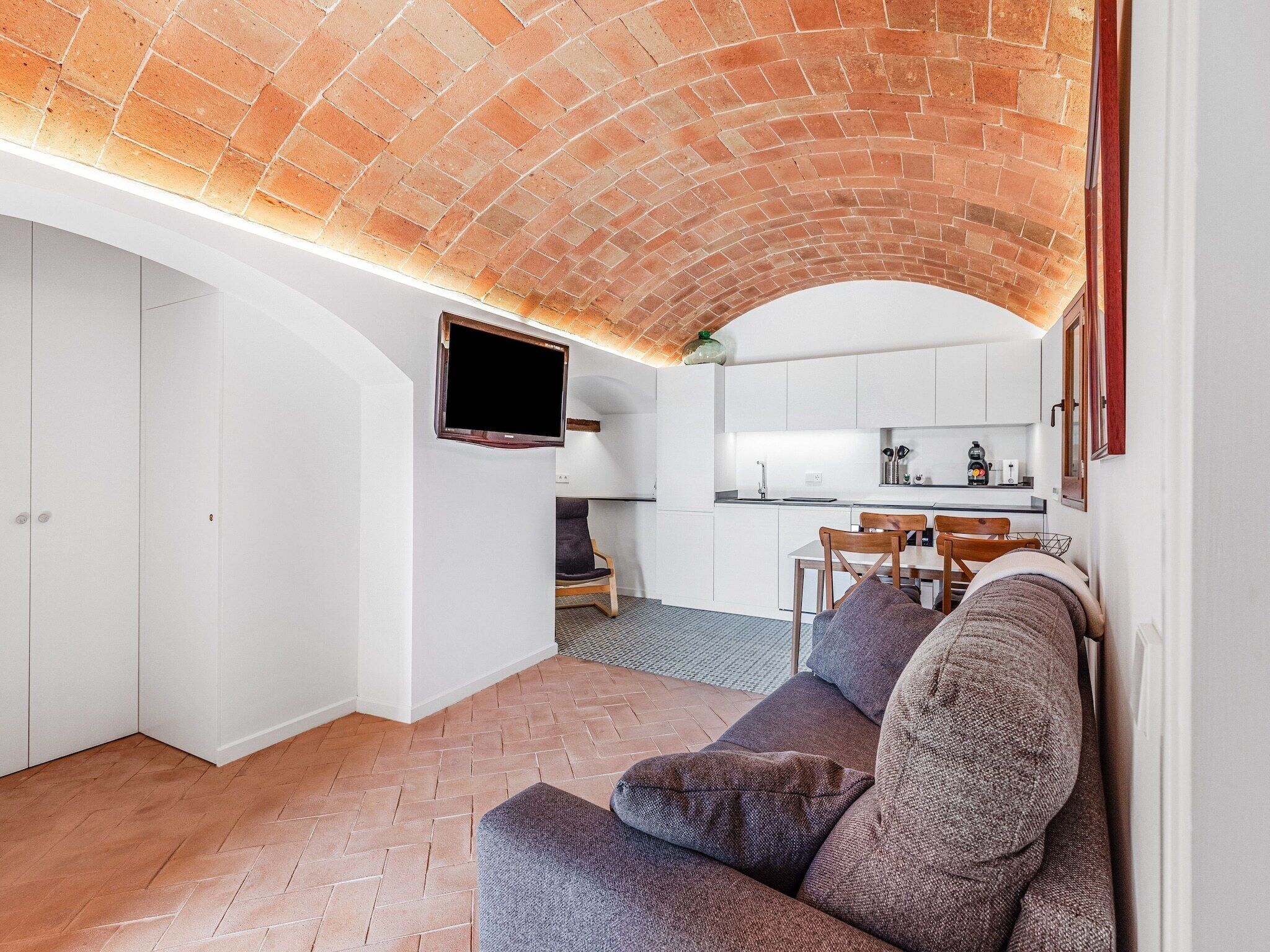 Un soggiorno moderno con soffitto in mattoni, una TV a schermo piatto montata a parete, un divano e un'area pranzo con una cucina sullo sfondo.
