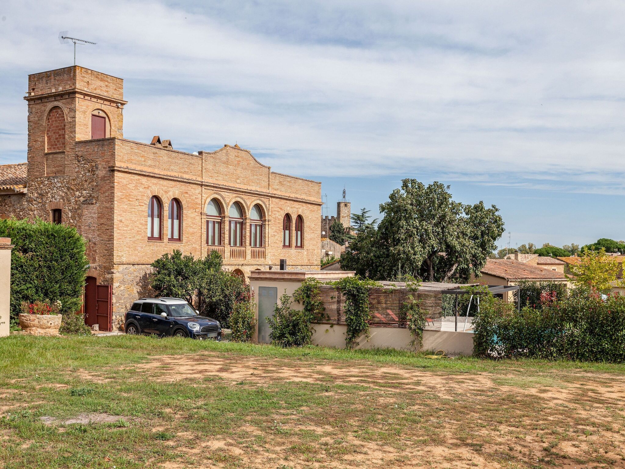 Un edificio histórico con una torre destacada y ventanas arqueadas, rodeado de vegetación y un coche estacionado.