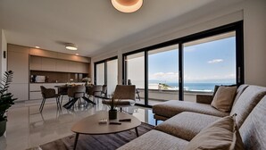 Una sala moderna con un sofá grande, una mesita de centro y una zona de comedor con sillas. Se aprecia una ventana grande con vistas al océano.