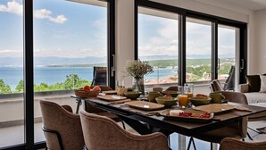 Un comedor con una mesa dispuesta para cuatro, ventanas amplias que ofrecen vistas al mar y a las montañas, y un florero con flores sobre la mesa.