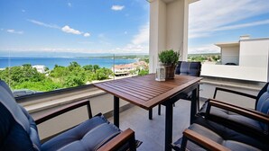 Un balcón con mesa y sillas de madera, una planta en maceta y vista al mar y la vegetación.
