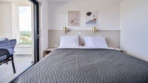 Habitación de hotel con una cama grande, dos mesitas de noche, una silla y un balcón con vistas.