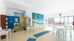 Una habitación luminosa y moderna con un ventanal grande, un mural de playa, un letrero de lavandería autoservicio y una zona de comedor con sillas y mesas.