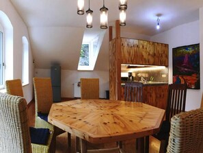 Un coin repas avec une grande table en bois entourée de chaises en osier, une cuisine à l’arrière-plan et un plafond en pente agrémenté de lucarnes.
