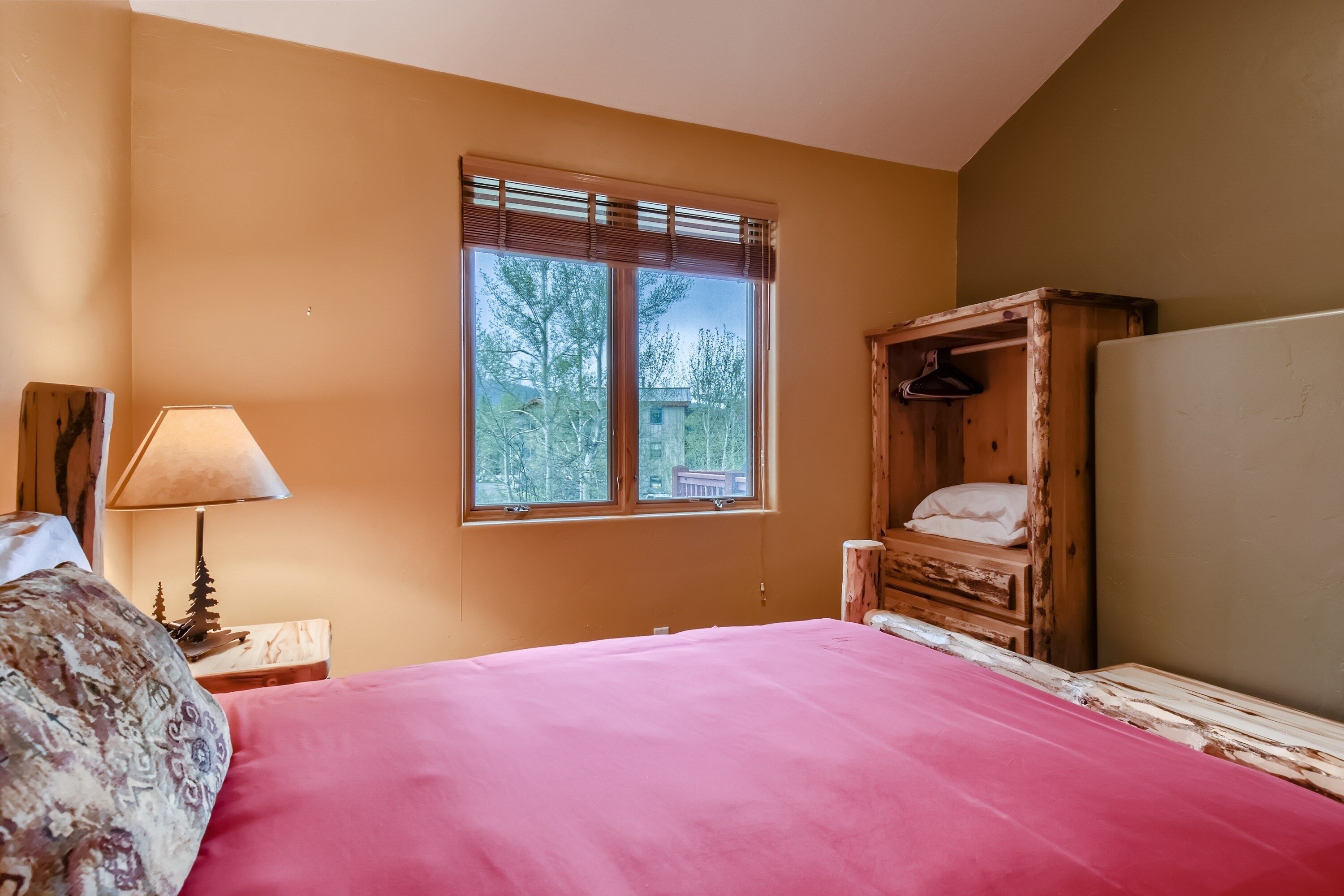 Ein Schlafzimmer mit großem Fenster, einem Bett mit rosa Bettdecke, einem Nachttisch aus Holz mit Lampe und einem Holzschrank.