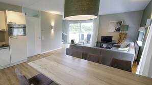 A modern kitchen with built-in appliances, a dining area with a wooden table and chairs, a living area with a sofa and TV, and a large window offering a view of the outdoors. - Nature Escape in Heimbach (Heimbach)