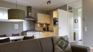 A modern kitchen with white cabinets, a stove, and a coffee maker. There is a dining area with a table and chairs, and a living room with a sofa and a decorative pillow. - Nature Escape in Heimbach (Heimbach)