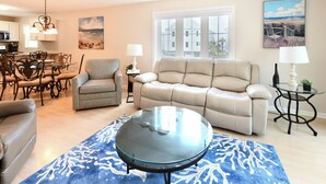 Een woonkamer met een glazen salontafel, een beige bank en een blauw vloerkleed met witte koraalmotieven.