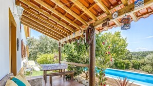 Uma área de estar coberta ao ar livre, vista para a piscina, vigas de madeira e objetos decorativos.