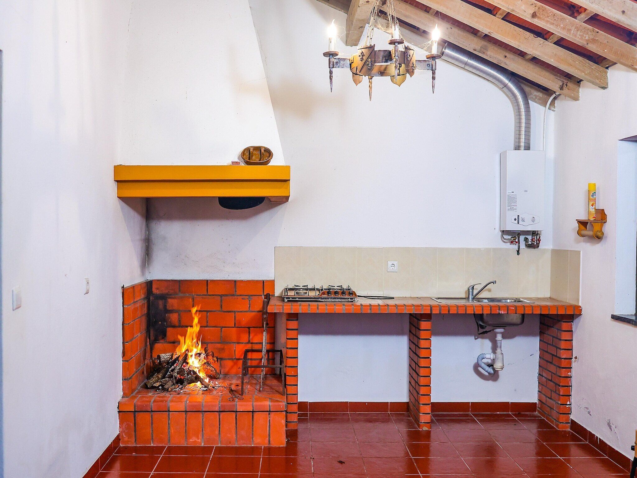 A kitchen with a brick fireplace, a gas stove, a hanging light fixture, and a white wall with a yellow shelf.