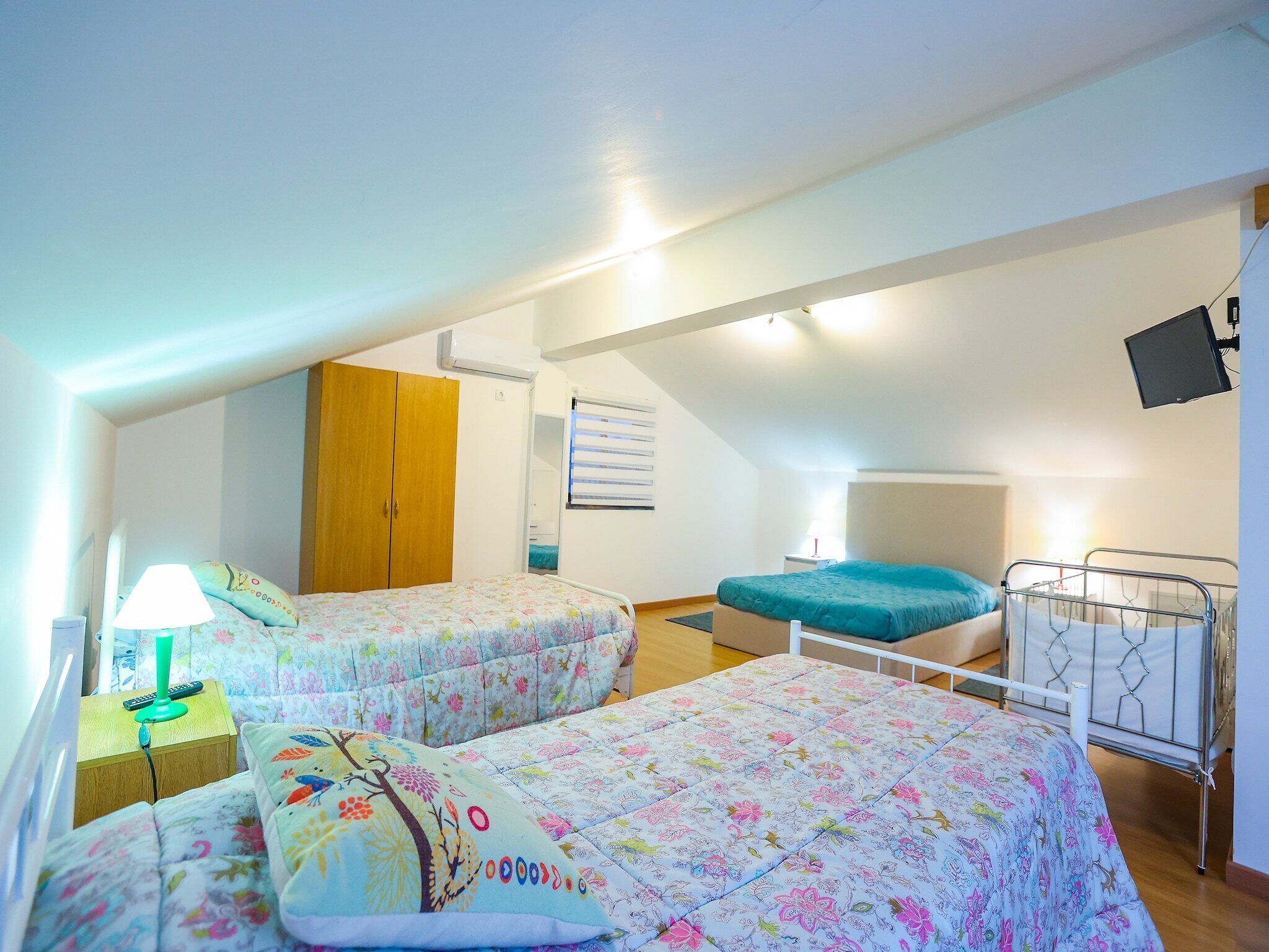A room with two beds, a wardrobe, a desk with a lamp, and a television mounted on the wall.