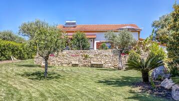 A house with a stone wall and a tiled roof, surrounded by a well-maintained lawn and various trees.