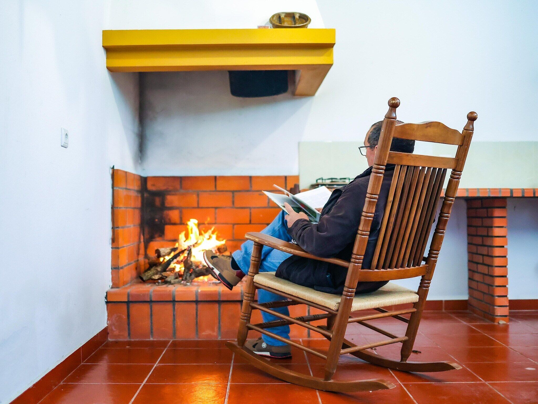 A person sitting on a rocking chair by a fireplace with a yellow shelf above it.