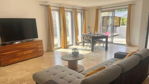 Una sala de estar moderna con un sofá modular grande, un televisor de pantalla plana sobre un mueble de madera y un comedor con mesa y sillas.