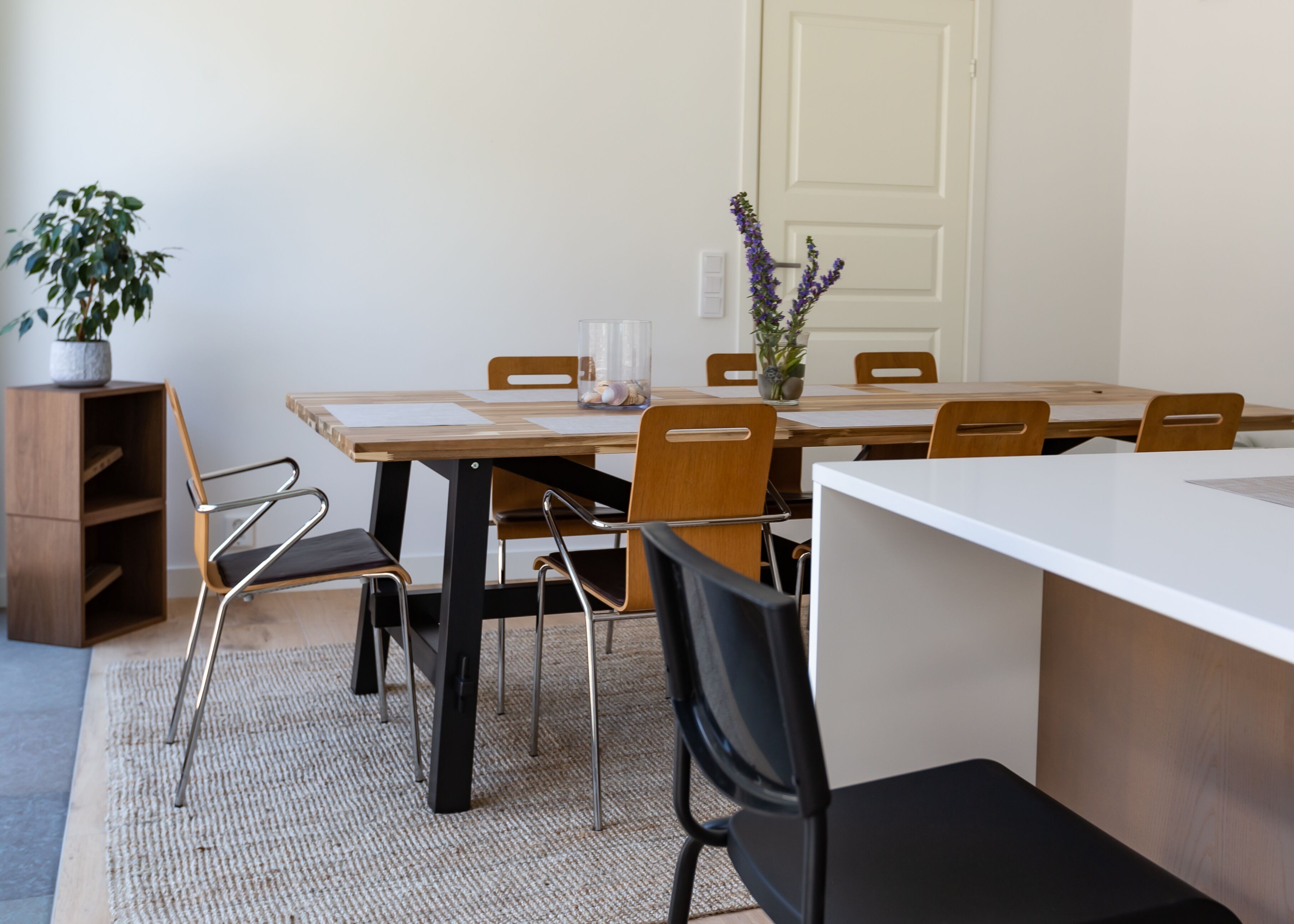 A dining area with a wooden table, six chairs, a potted plant, and a vase with flowers.