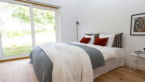 A modern bedroom with a large bed, a bedside table, a floor lamp, and a window with a view of greenery. - Halltorp Townhouse (Borgholm)