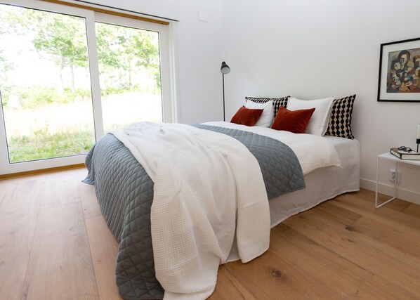 Un dormitorio moderno con una cama grande, una mesita de noche, una lámpara de pie y una ventana con vistas a la vegetación.