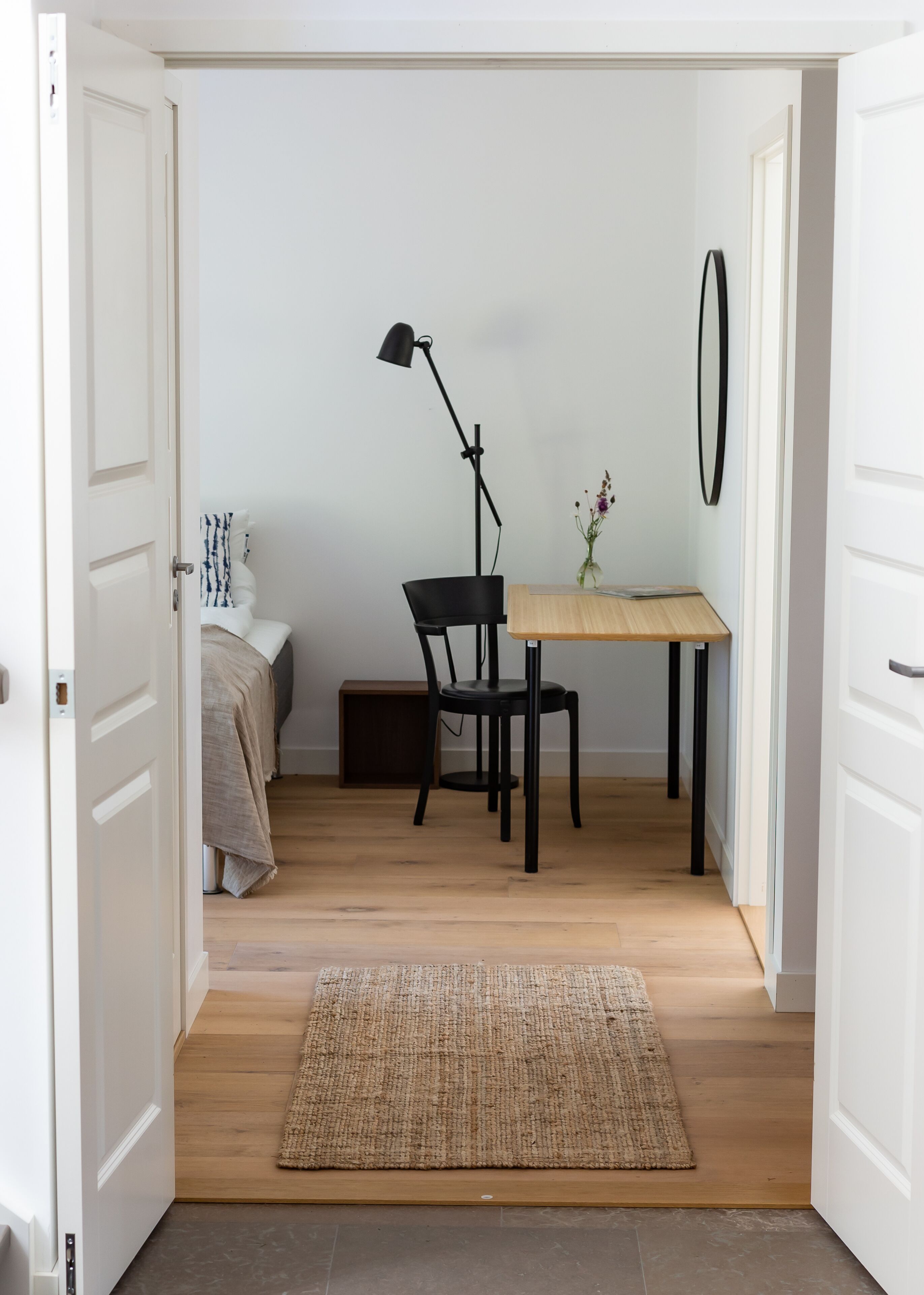 A room with a wooden floor, a small table with a chair, a lamp, and a vase with flowers.